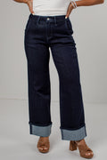 A person wearing dark indigo wide-leg jeans with rolled cuffs, paired with a white top and brown heeled sandals, shown from the waist down against a light gray studio background.