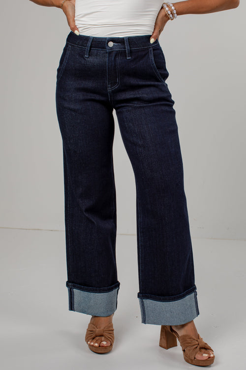 A person wearing dark indigo wide-leg jeans with rolled cuffs, paired with a white top and brown heeled sandals, shown from the waist down against a light gray studio background.