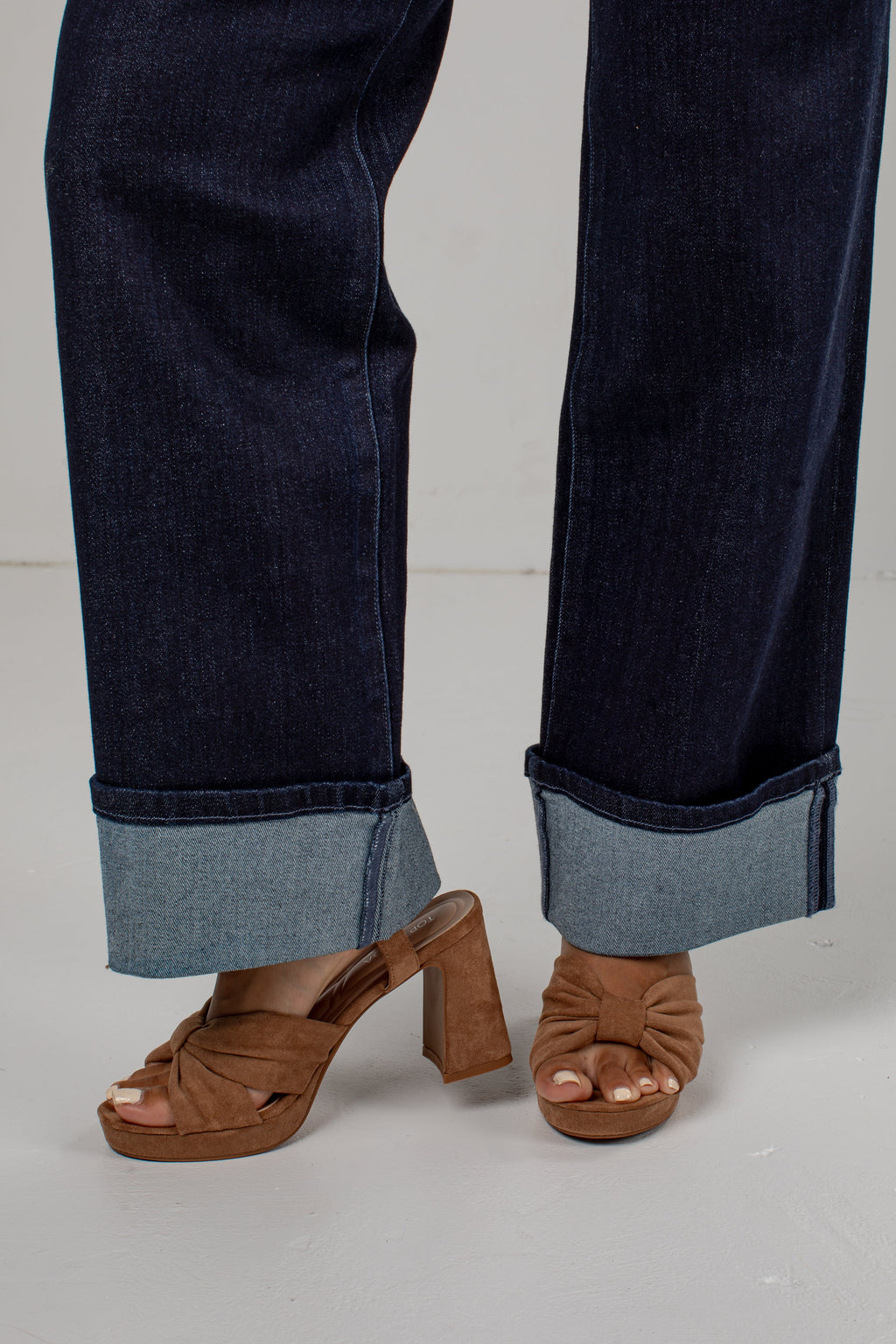 A close-up of a person's legs wearing dark blue cuffed jeans and brown block-heel mule sandals against a light gray studio background.