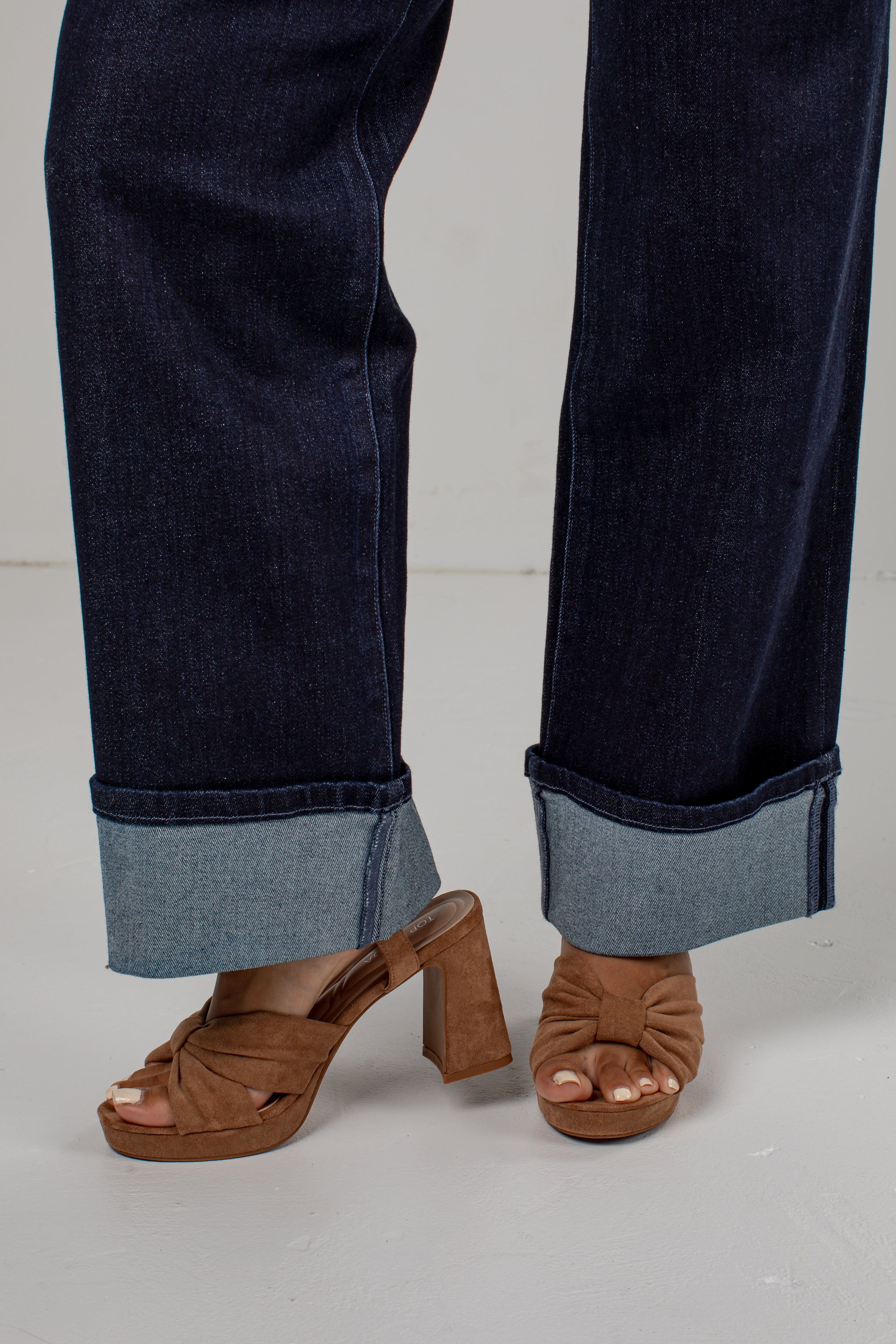 A close-up of a person's legs wearing dark blue cuffed jeans and brown block-heel mule sandals against a light gray studio background.